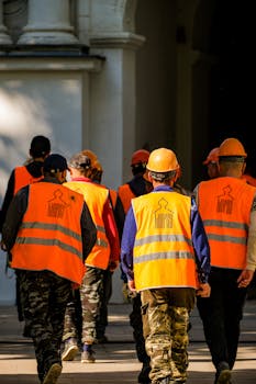 Group of construction workers wearing orange vests and helmets walking in urban area.