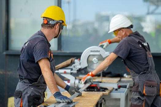 Two construction workers cutting a metal beam with a circular saw, wearing safety gear on site.